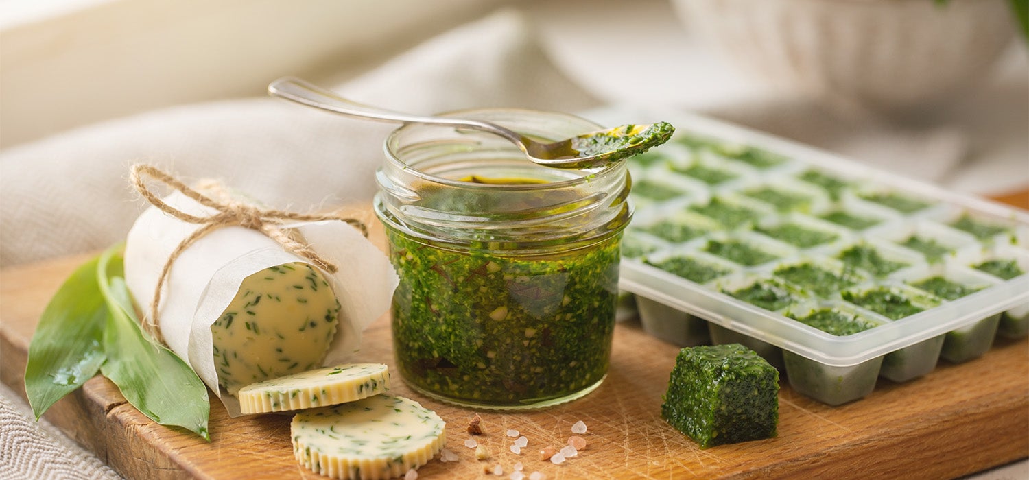 Ein Glas Bärlauchpesto, eine Rolle Bärlauchbutter und eine Eiswürfelform gefüllt mit grünem Bärlauchpüree auf einem Holzbrett.