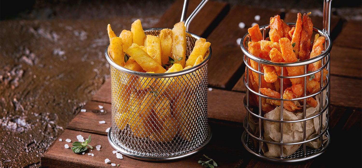 Zwei Metallkörbe mit Kartoffel- Pommes und Süßkartoffel-Pommes, serviert auf einem Holzbrett