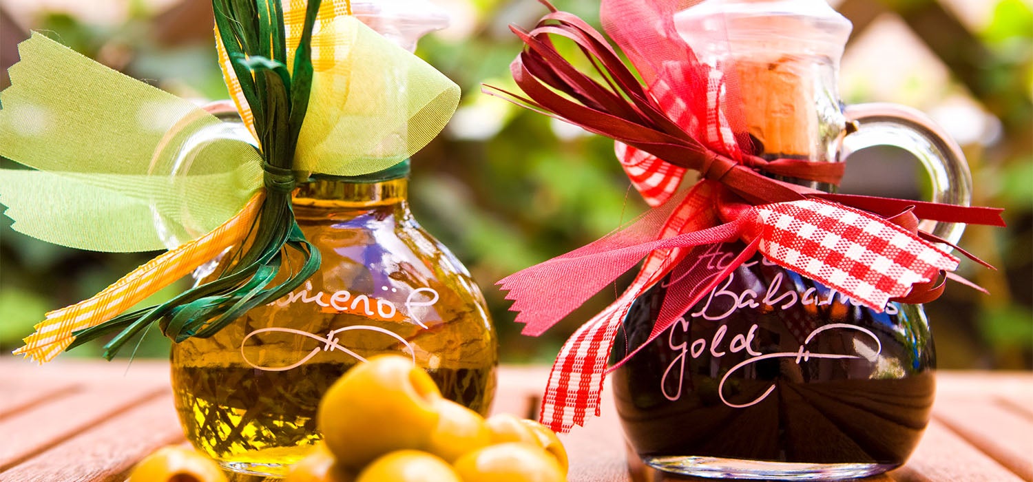 Zwei verzierte Flaschen mit Olivenöl und Balsamico auf einem Holztisch, umgeben von grünen Oliven