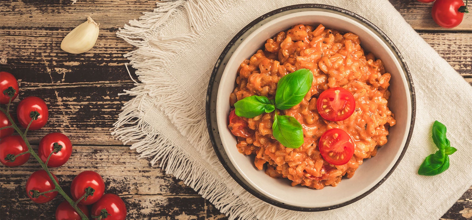 Ein Teller Tomaten-Risotto steht auf einem Holztisch mit Knoblauch und frischen Tomaten daneben