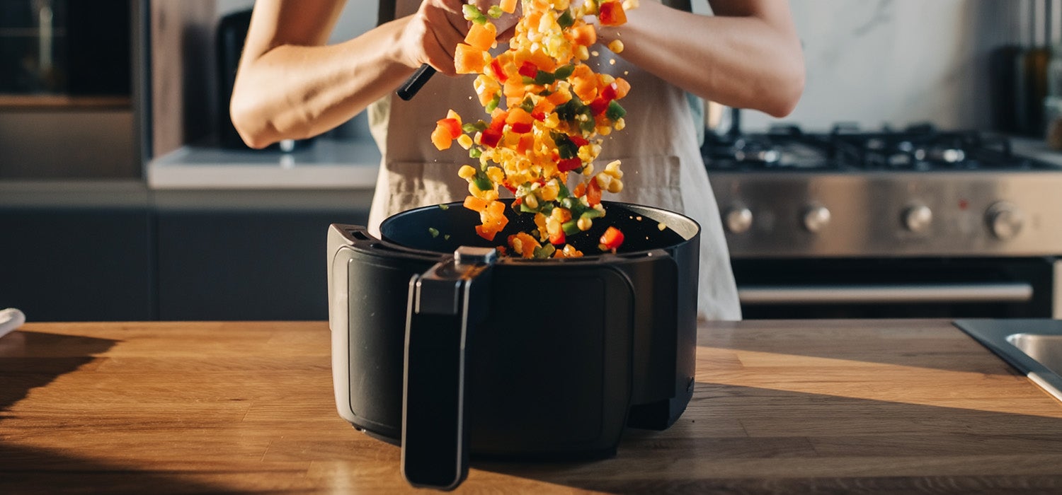 Eine Person schüttelt den Garkorb einer Heißluftfritteuse, in dem sich buntes Gemüse befindet, um es zu wenden