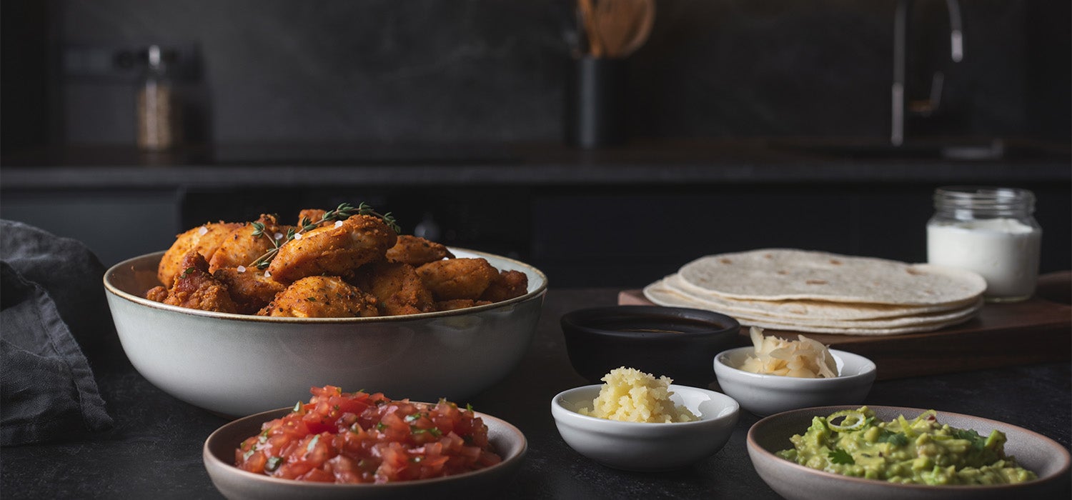 Fertig gebratenes, gewürztes Hähnchenfleisch in einer Schale, daneben liegen verschiedene internationale Zutaten wie Sojasauce, Ingwer, Joghurt, Knoblauch, weiche Tortilla-Wraps und Schalen mit Salsa und Guacamole.]