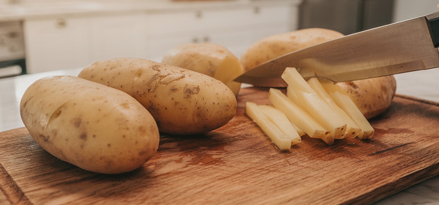 Rohe Kartoffeln liegen nebeneinander auf einem Holzbrett, eine davon wird gerade in Stifte geschnitten.]