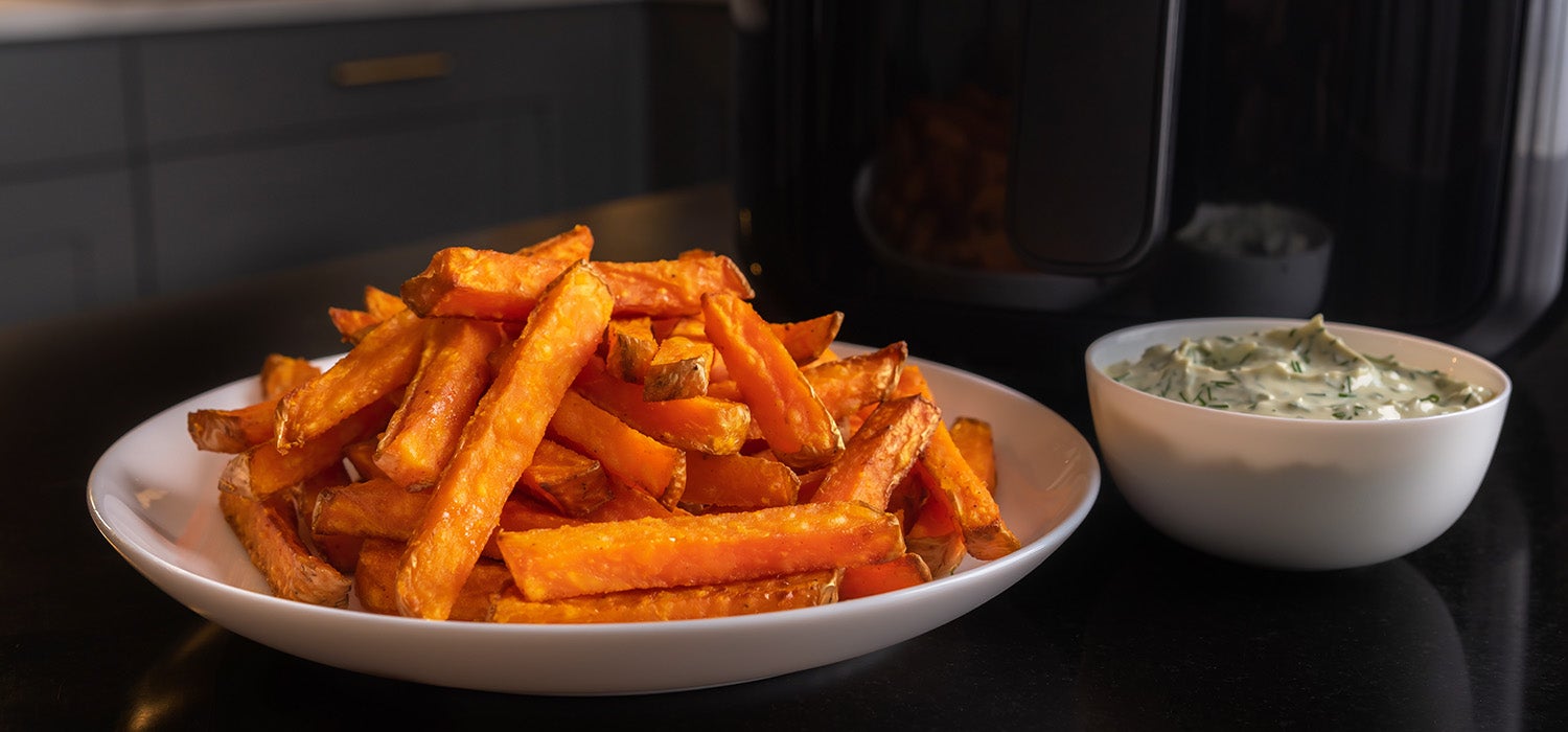 Ein einfacher, heller Teller, auf dem frisch zubereitete, leuchtend orange Karotten-Pommes aus dem Airfryer liegen. Daneben ein kleines Schälchen mit Kräuterquark. Der Fokus liegt klar auf den knusprig aussehenden Karotten-Pommes