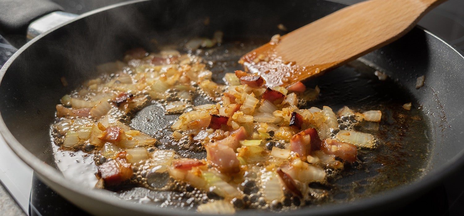 Nahaufnahme von Zwiebel- und Knoblauchwürfeln und Speckwürfeln, die in einer Pfanne goldbraun anbraten