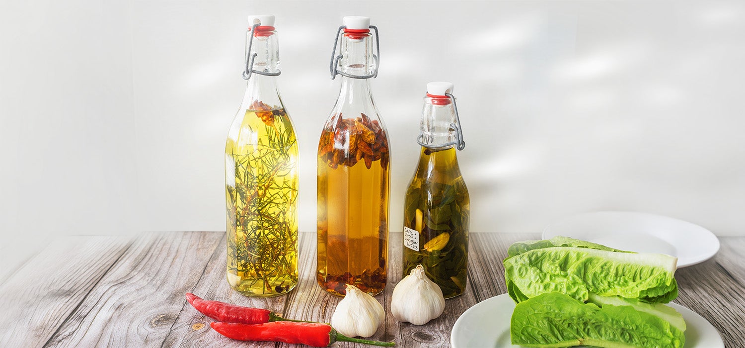 Drei transparente Glasflaschen mit Bügelverschluss stehen auf einem Holztisch und sind mit aromatisiertem Öl gefüllt, in dem Kräuter, getrocknete Chilischoten und Knoblauchzehen sichtbar eingelegt sind, daneben liegen frische rote Chilis, Knoblauchknollen und grüne Salatblätter vor hellem, natürlichem Hintergrundlicht.