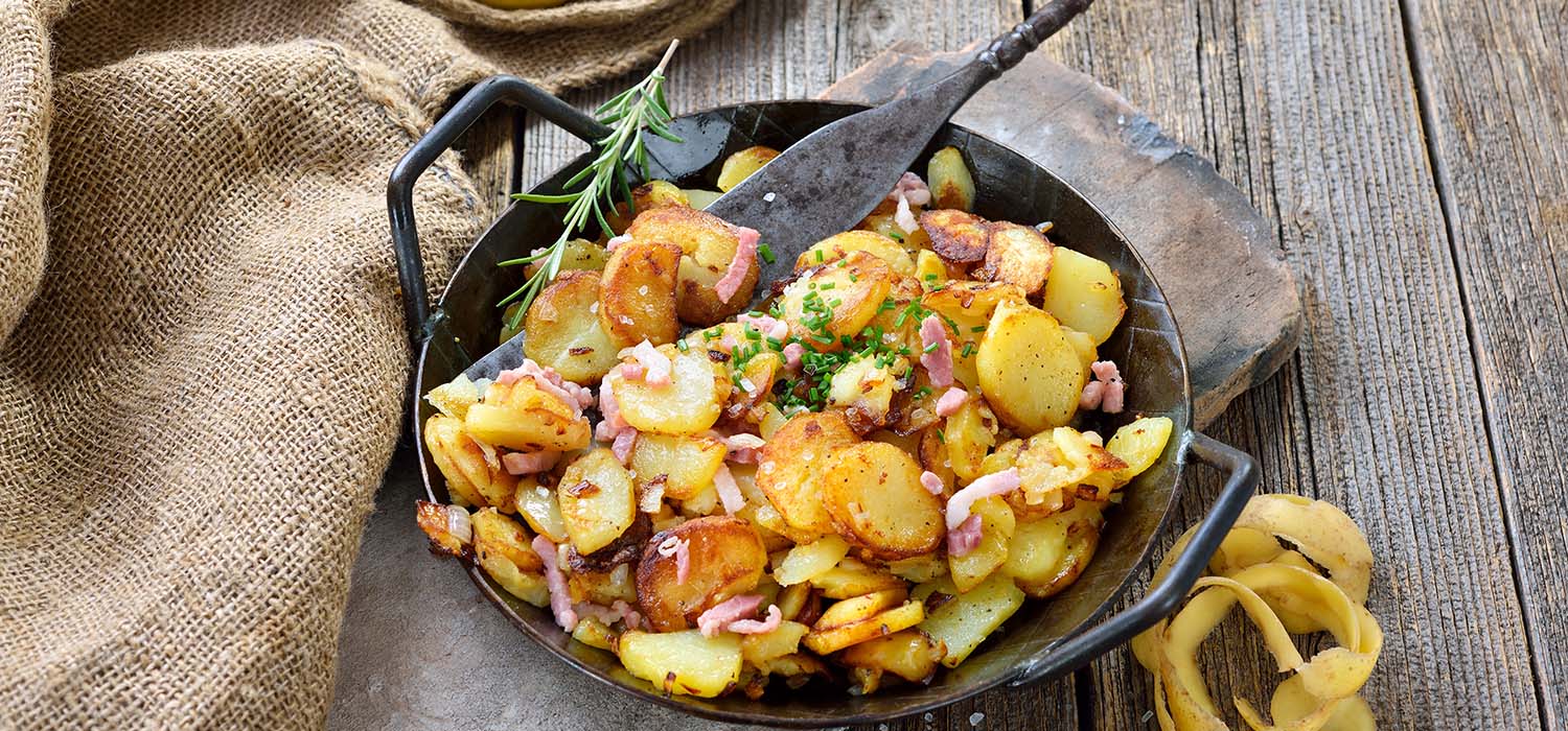 Bratkartoffeln mit Speck und Kräutern angerichtet in einer Pfanne auf einem Holzbrett.
