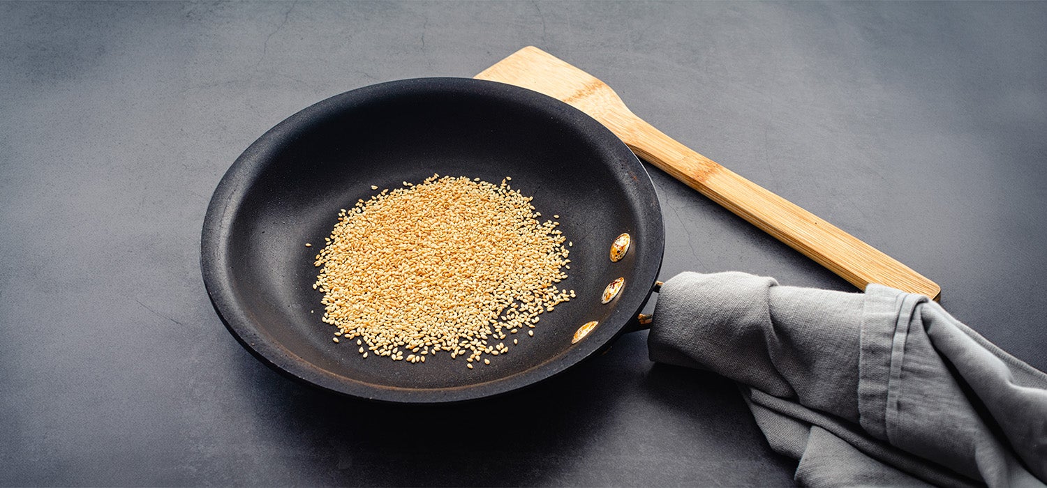 Sesam in der Pfanne, daneben ein Kochlöffel.