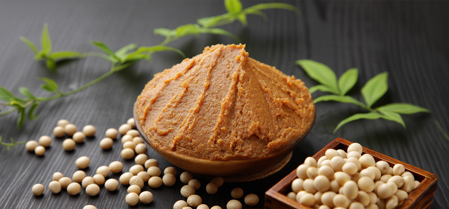  Braune Miso-Paste schön angerichtet in einer schwarzen Schüssel, daneben Sojabohnen.