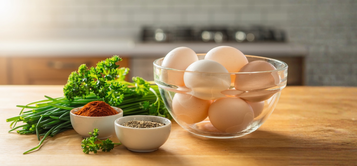 Eine bunte Mischung aus frischen Kräutern (Schnittlauch, Petersilie) und kleinen Schälchen mit Gewürzen (Paprika, Pfeffer) neben einer Schüssel mit rohen Eiern