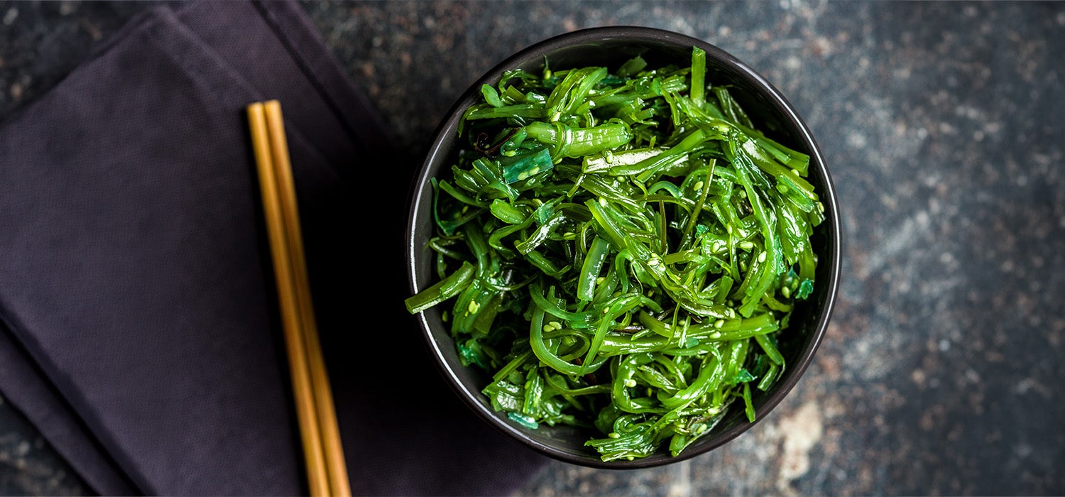  Eine schwarze Schale ist mit einem grünen Algen-Salat aus glänzenden, in Streifen geschnittenen Meeresalgen gefüllt, die leicht mit Sesamsamen bestreut sind und auf einer dunklen Steinoberfläche neben Holzstäbchen angerichtet sind.
