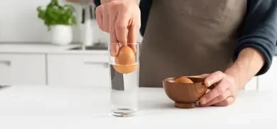 Eine Hand hält ein Ei über ein Glas Wasser, um den Frische-Test zu demonstrieren