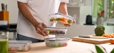 Person stapelt Meal-Prep-Boxen mit Gemuese auf Kuechentisch neben Schneidebrett mit Brokkoli und Karotten