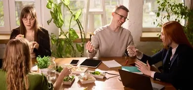 Mehrere Personen an Tisch mit Takeaway-Boxen, Laptops, Notizbuch, Glaesern und Pflanzen im Hintergrund