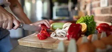 Hand schneidet rote paprika auf schneidebrett, daneben pilze, zitrone, salat, eier und tomaten