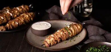 Kartoffelspiralen am Spieß mit Feta und Kräutern auf Teller, daneben Schale mit Dip, Hand streut Kräuter