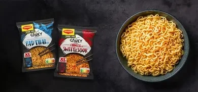 Zwei Maggi Instantnudel-Packungen Pad Thai und Sweet & Sour neben Schüssel mit gekochten Nudeln auf dunklem Hintergrund