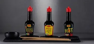 Drei Maggi Würze Flaschen mit rotem Verschluss auf Schieferplatte vor grauem Hintergrund
