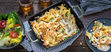 Auflaufform mit gebackenen Nudeln, Gemuese und Kaese, daneben Tellerportion und Salat auf Holztisch