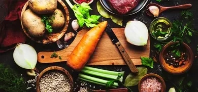Karotte, Kartoffeln, Zwiebel, Sellerie, Rindfleisch, Knoblauch, Kraeuter und Gewuerze auf Schneidebrett in Draufsicht
