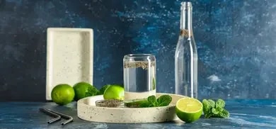 Glas mit Chia-Wasser, Limetten, Minze und Flasche auf Tablett vor blauem Hintergrund