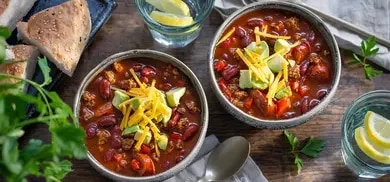 Zwei Schalen Chili con Carne mit Kidneybohnen, Avocado und Käse auf Holztisch.