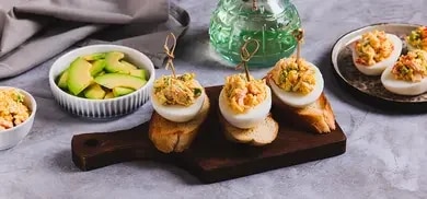 Gefüllte Eierhälften auf Baguettescheiben mit Spießen, Avocadoscheiben und Schüsseln auf Tisch angerichtet