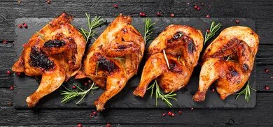 Vier gegrillte Hähnchenschenkel mit Grillspuren und Kräutern auf dunkler Platte angerichtet