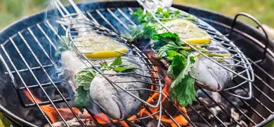 Fisch im Grillkorb mit Zitronenscheiben und Kraeutern ueber Holzkohlegrill mit Flammen und Rauch