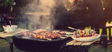 Grill mit Spießen aus Fleisch und Gemüse sowie Würstchen, Teller mit bunten Spießen daneben, Rauch über Grill