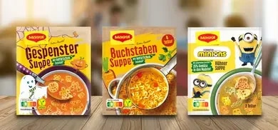 Drei Maggi Suppenpackungen mit Minions Huehnersuppe, Gespenstersuppe und Buchstabensuppe auf Holztisch