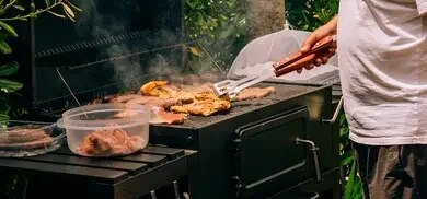 Person grillt Fleisch und Würstchen auf Holzkohlegrill, daneben Schalen mit rohem Fleisch und Grillzange