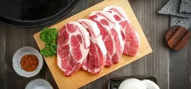 Mehrere rohe Schweinenackensteaks auf Holzbrett, daneben Petersilie und Schalen mit Gewürzen