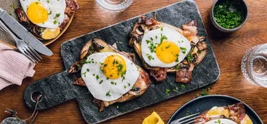 Offene Brote mit Spiegeleiern, Speck und Pilzen auf Schieferplatte, mit Schnittlauch bestreut