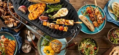 Grill mit Gemüse, Spießen und Käse, umgeben von Schalen mit Salaten und Brot auf Holztisch