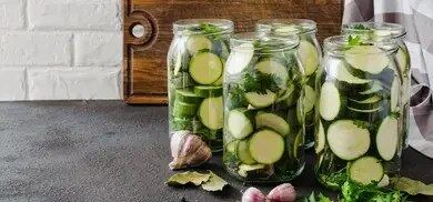 Vier Glaeser mit Zucchinischeiben, Knoblauch, Lorbeerblaettern und Kraeutern auf Arbeitsflaeche