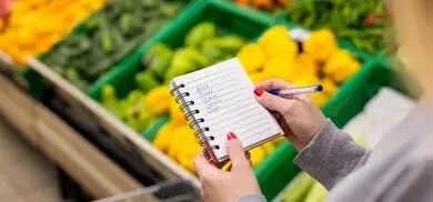 Hand mit Notizblock und Einkaufsliste vor Gemüseregal im Supermarkt