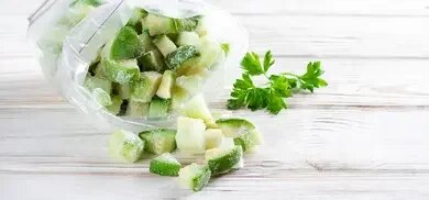 Gefrorene Zucchiniwürfel in offenem Gefrierbeutel auf heller Holzoberfläche mit Petersilie