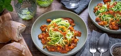 Zucchininudeln mit Tofu-Bolognese auf Tellern, dazu Brot und Besteck auf Holztisch