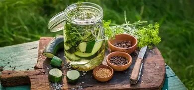 Einmachglas mit Zucchinischeiben, Dill, Knoblauch und Gewuerzen auf Holzbrett im Freien