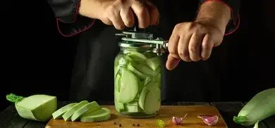 Haende verschliessen Einmachglas mit Zucchinischeiben auf Holzbrett, daneben Knoblauch und Zucchini