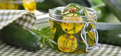 Einmachglas mit Zucchinischeiben in Oel, Rosmarin und Pfefferkoernern neben frischen Zucchini