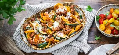 Zucchini-Moussaka mit Hackfleisch und geschmolzenem Käse in ovaler Auflaufform mit Tomatensalat im Hintergrund