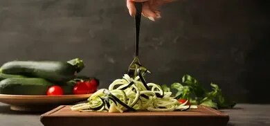 Zucchininudeln auf Schneidebrett mit Gabel, ganze Zucchini und Tomaten im Hintergrund