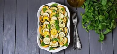 Servierplatte mit Zucchinischeiben, Minzblaettern und Besteck auf grauer Holzoberflaeche