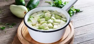 Zucchiniwürfel in Topf mit Wasser auf Holzbrett, daneben ganze Zucchini und Petersilie