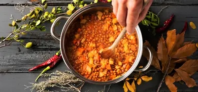 Kochtopf mit Linseneintopf, Gemüse und Holzlöffel, umgeben von Kräutern und getrockneten Chilis
