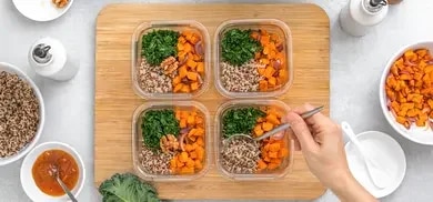 Vier Glasboxen mit Quinoa, Grünkohl und Süßkartoffeln auf Holzbrett, Hand mit Löffel beim Befüllen