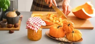 Glas mit eingelegten Kürbiswürfeln vor geschnittenem Kürbis, kleinen Kürbissen und Kürbiskernen
