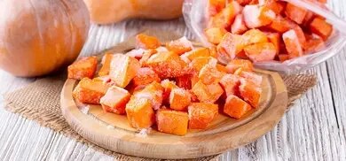 Gefrorene Kürbiswürfel auf einem Holzbrett neben Kürbissen und einem Gefrierbeutel